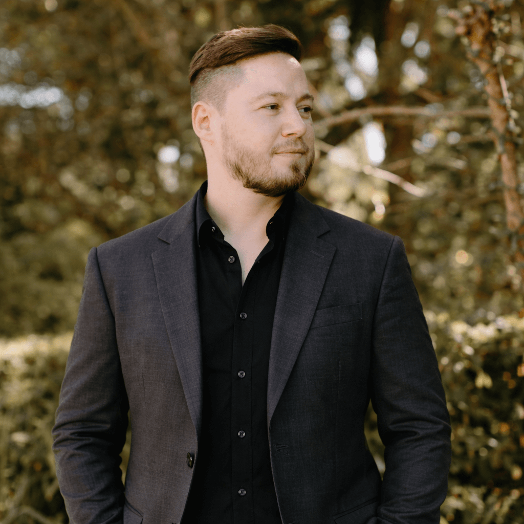 Photograph of Lee with tidy modern hair cut, wearing a dark coloured suit, with tropical plants in the background.