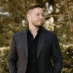 Photograph of Lee with tidy modern hair cut, wearing a dark coloured suit, with tropical plants in the background.